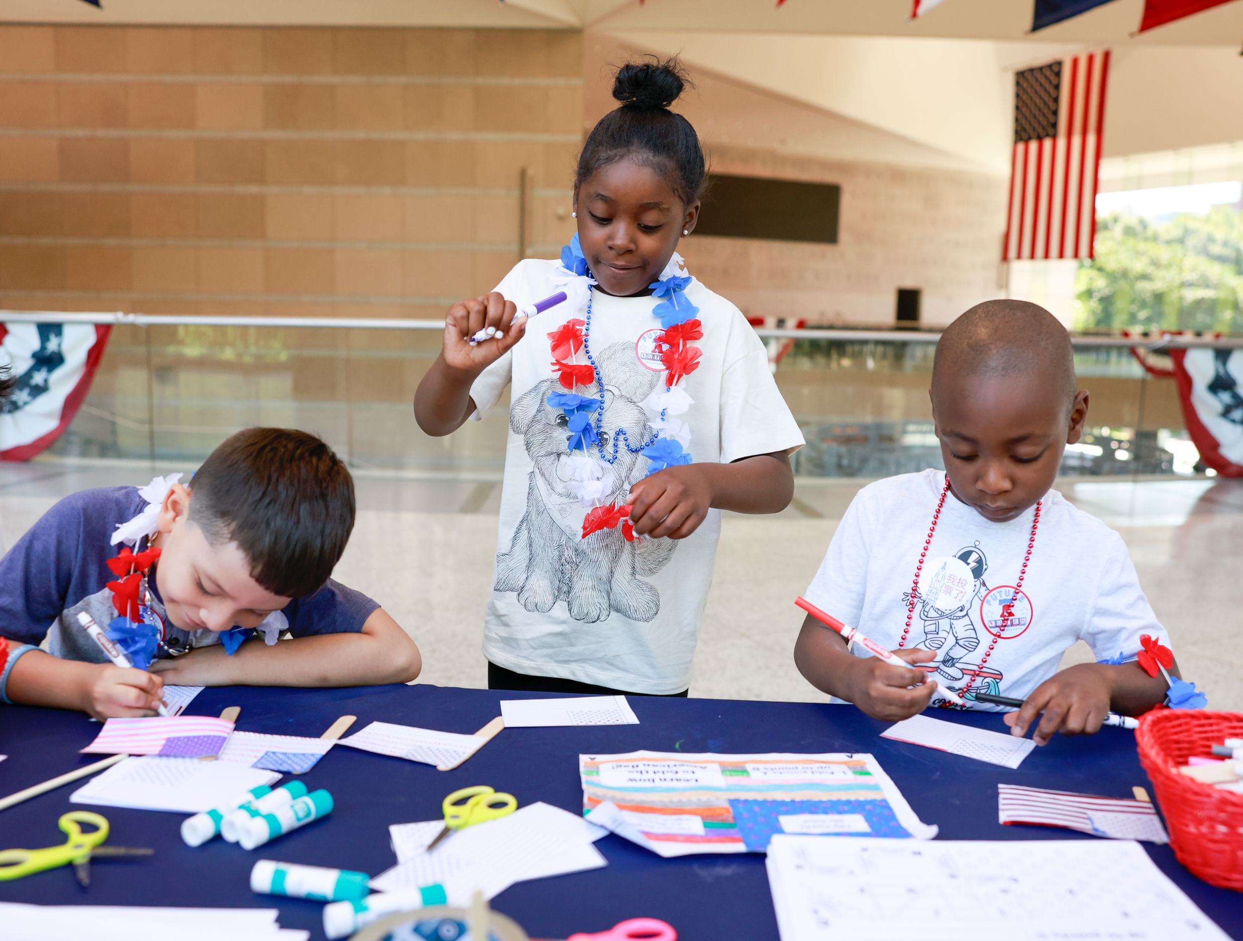 Three children wearing festive necklaces work happily with markers at an activities table with a blue cloth.