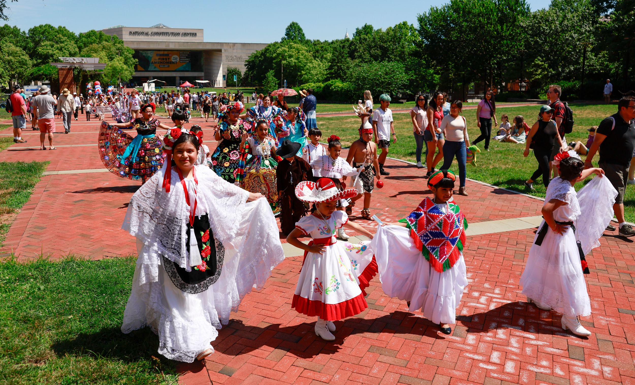 Girls in colorful, lacy, embroidered Central American dresses march together under a sunny blue sky on a red brick pathway.