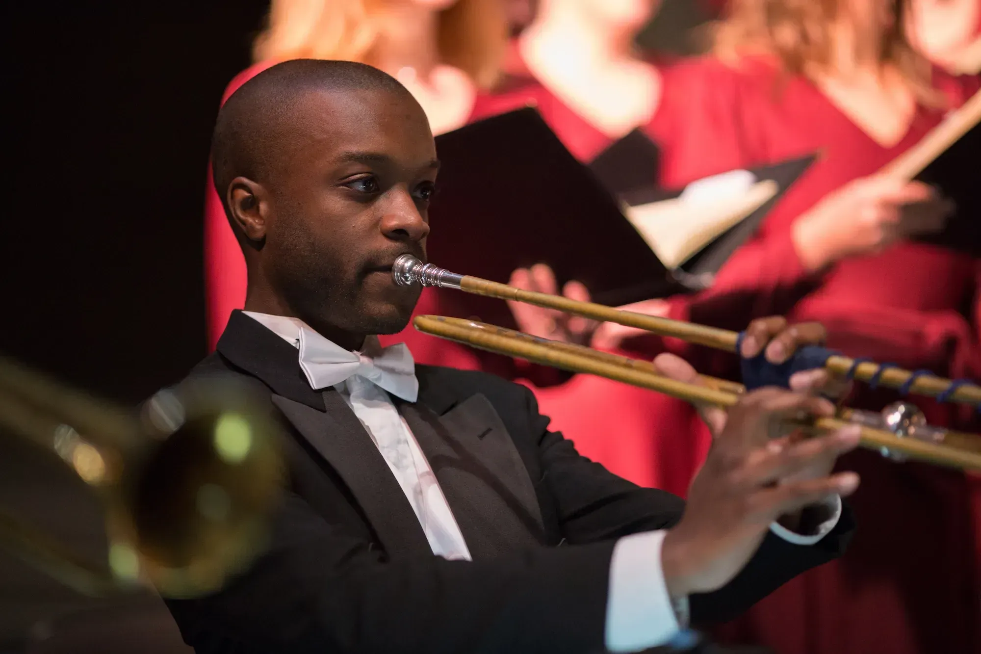 Close up of Bland, a Black man, in a black and white formal suit with bowtie plays the trumpet amongst other players.