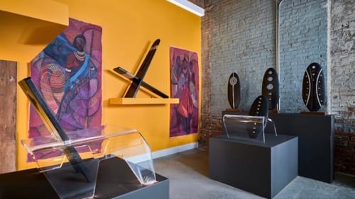 Gallery view with a birthing chair at left & black floor meeting a yellow wall with large colorful works with African figures