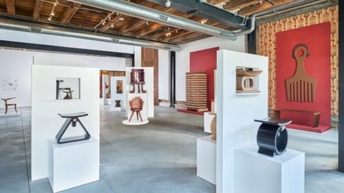 Gallery view with several of Tariku’s small, sleek wooden chairs & historic artifacts on modern white pedestals.