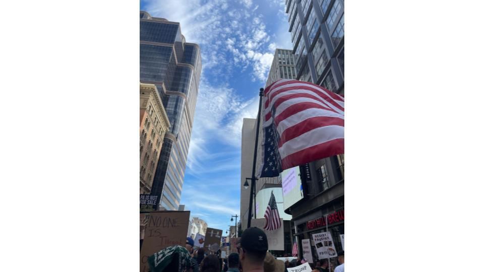 View over the top of a large protest crowd carrying flags and signs under a blue sky.