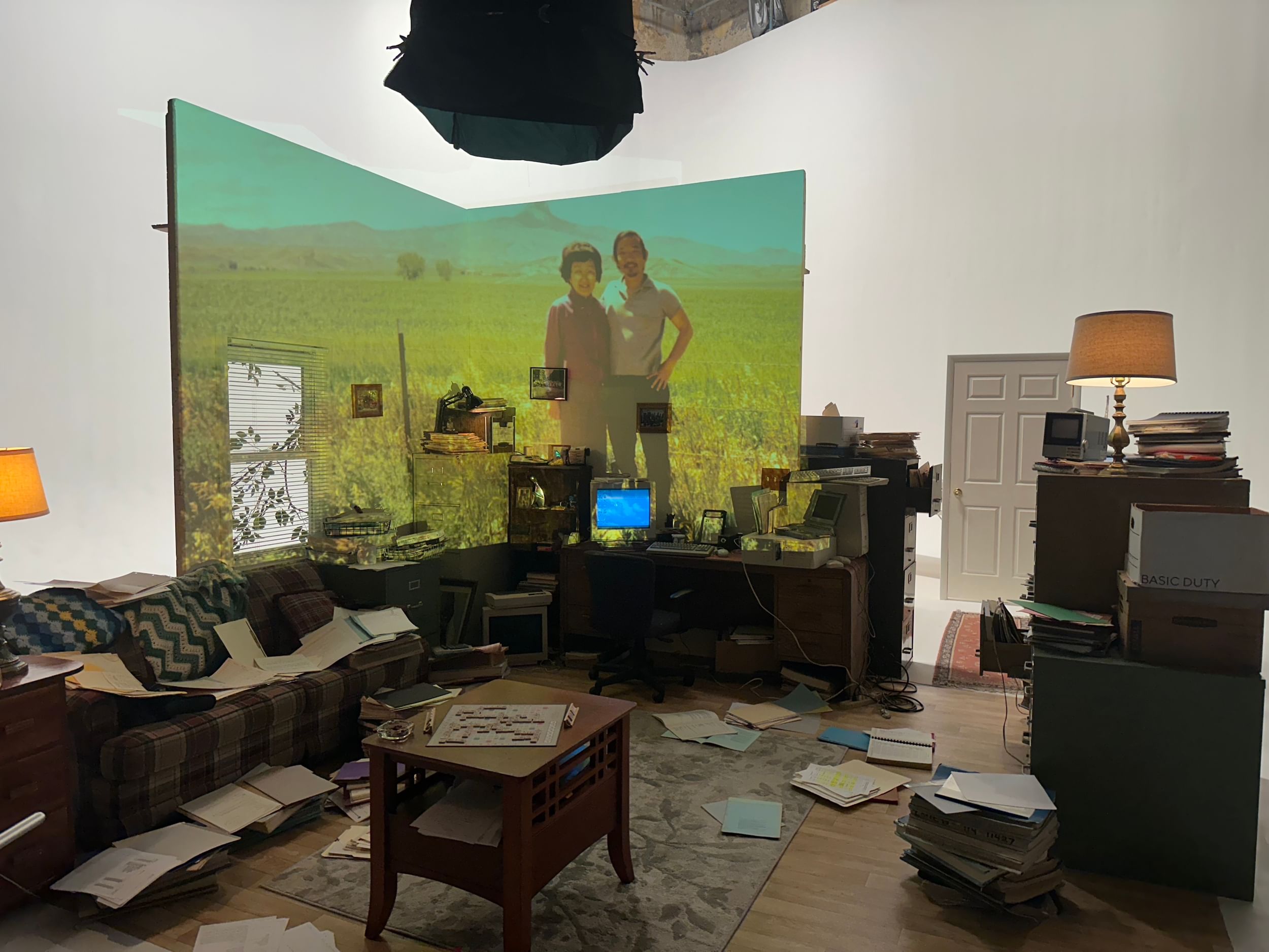 Inside a living room, paper scattered everywhere, and a large projection of an Asian man and woman lighting up one corner.