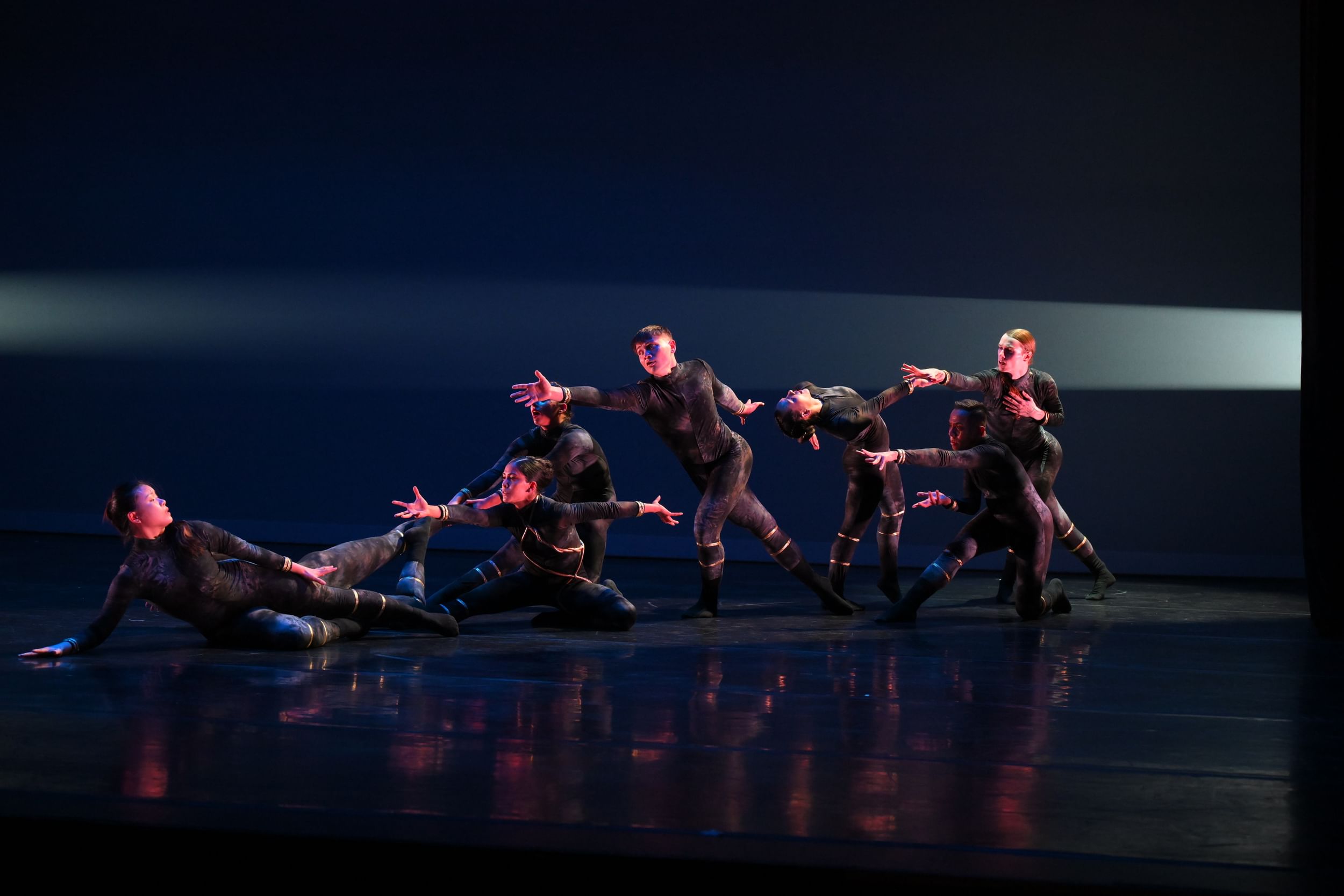 6 dancers in identical black & gold bodysuits reach dramatically in different poses toward 2 dancers on the floor at left.