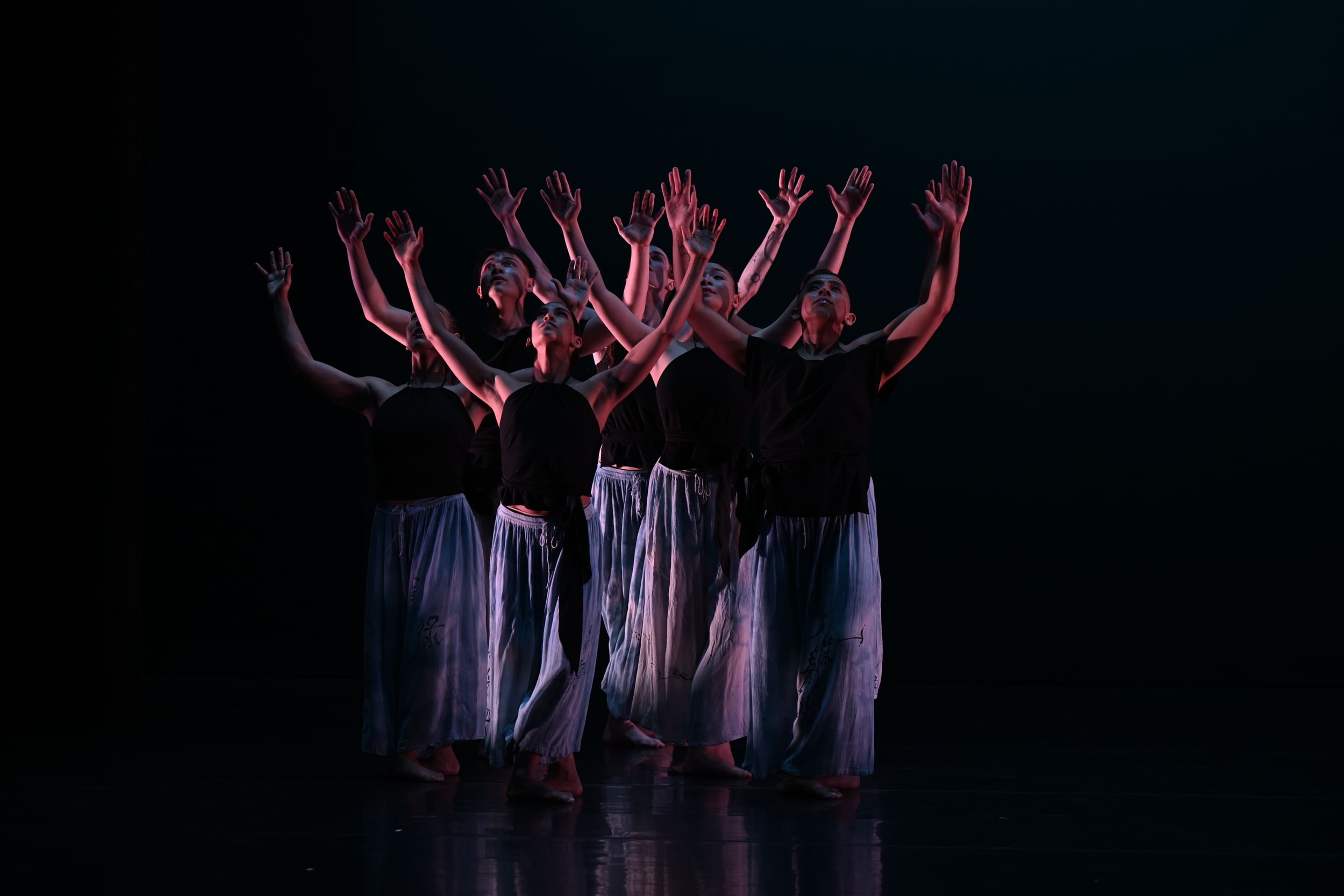 6 dancers in black tops and flowing blue pants spread their hands upward, making a overlapping forest of their arms.