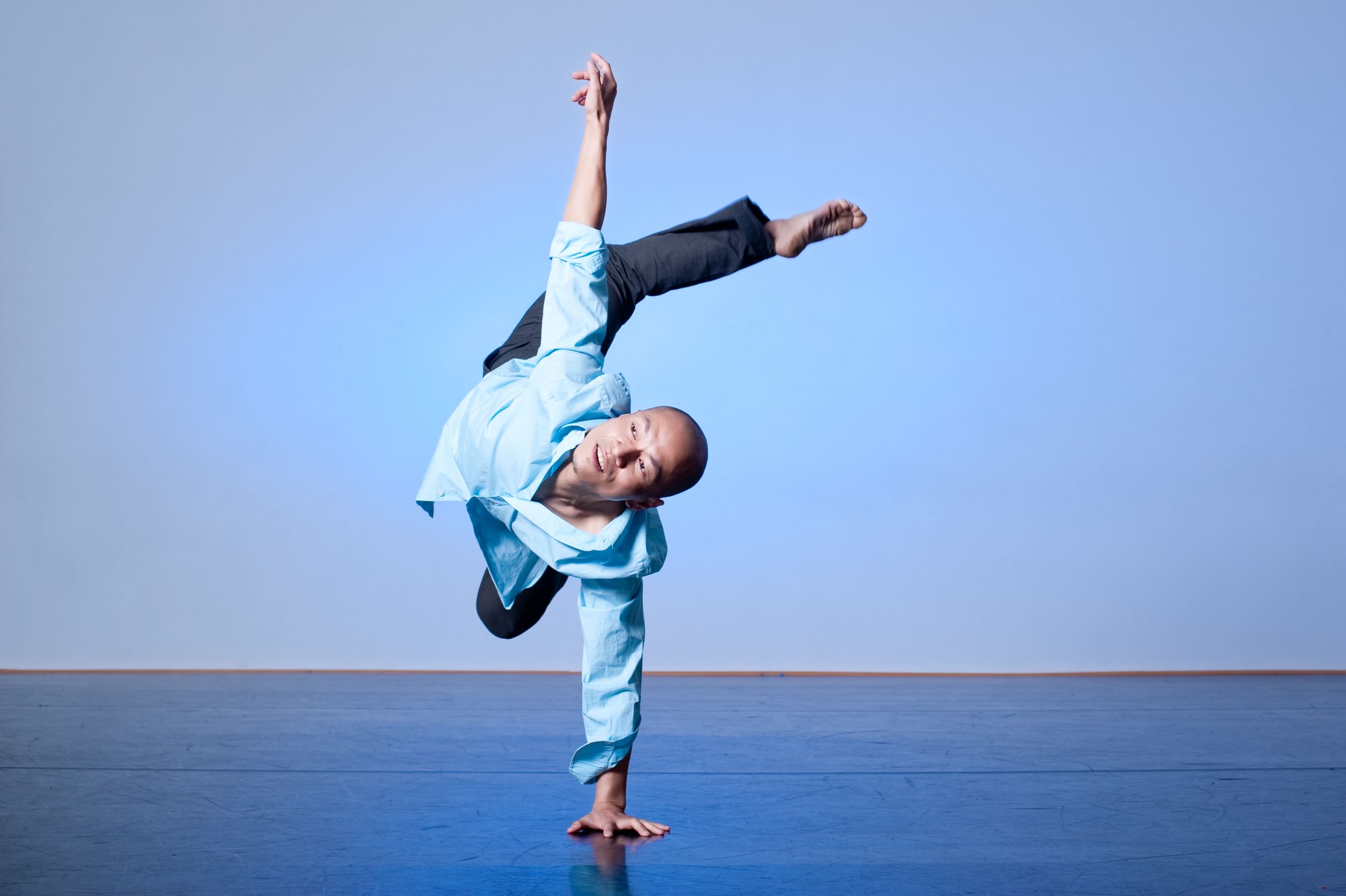 Lin, in a loose blue button-down, caught in a dynamic move with one hand on the blue-lit stage and all other limbs aloft.