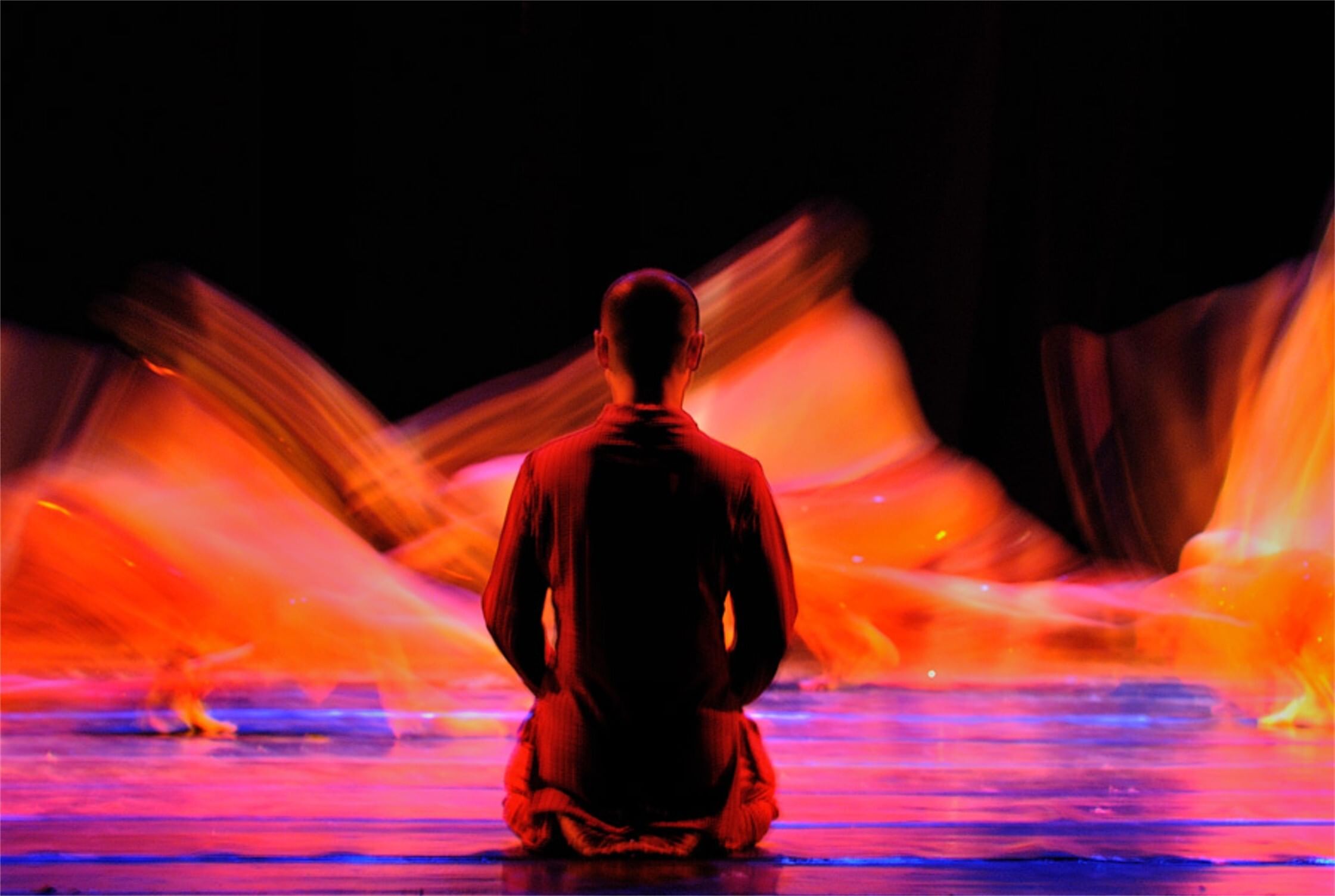 Seen from behind, Lin kneels while other dancers, a hot blur of orange limbs, cavort upstage.