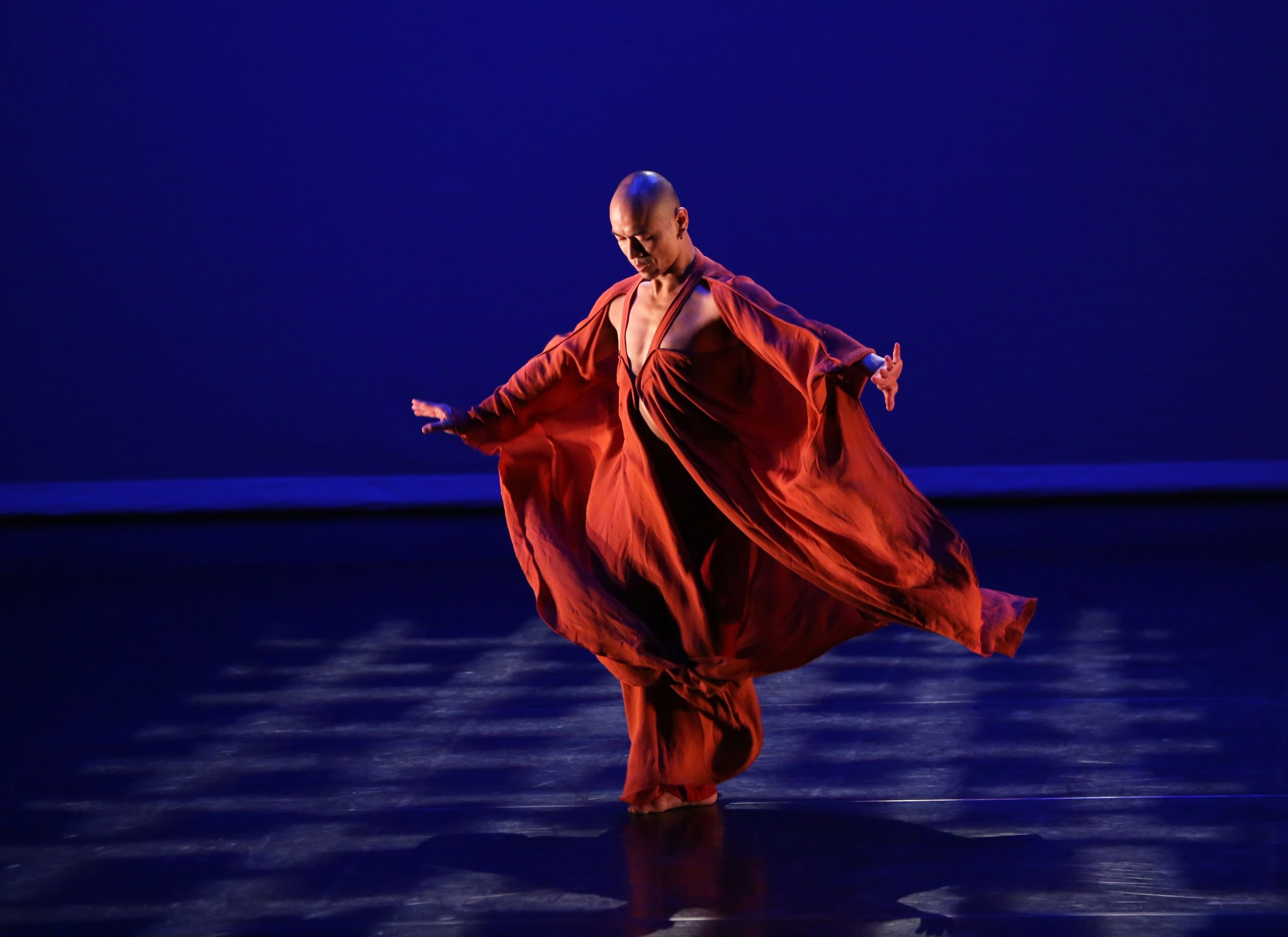 Lin, a bald male Asian dancer, stands with arms spreading downward, in a flowing orange garment rippling in wind.