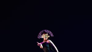 Ma, a single dancer in black on a dark stage, her face covered with a fan she holds in her mouth. She strikes an angular pose