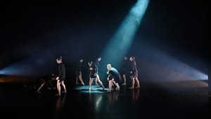 Small on a dark stage, 9 black-clad dancers move around a spotlight, which catches on in the middle clutching her chest.