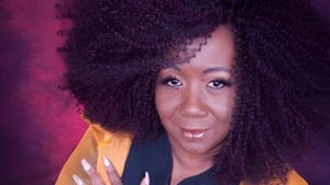Headshot of Moore, a smiling Black woman with luxuriant hair and an orange jacket, crossing her arms on her chest.