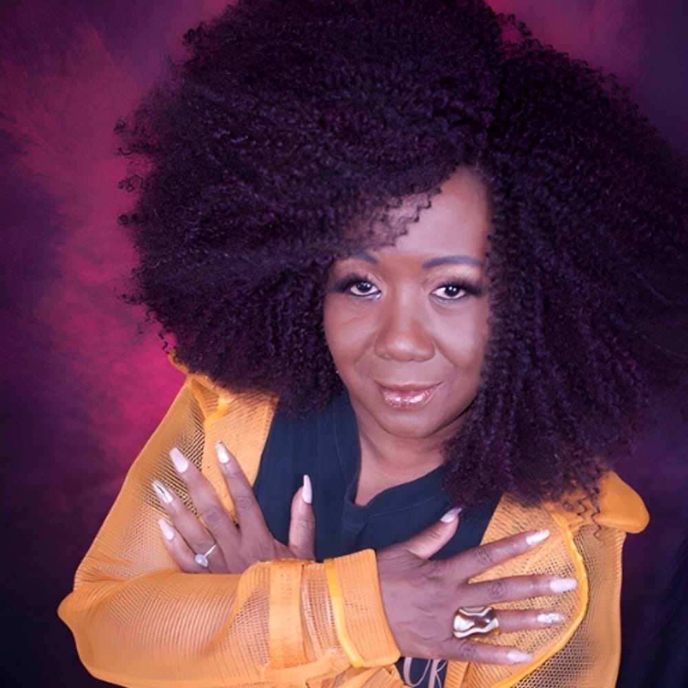 Headshot of Moore, a smiling Black woman with luxuriant hair and an orange jacket, crossing her arms on her chest.