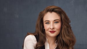 Bailey, a white woman with luxuriant wavy brown hair, poses in a white shirt and black vest.