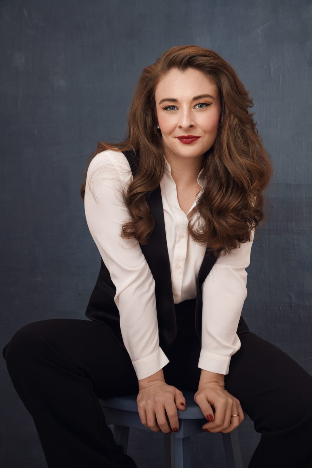 Bailey, a white woman with luxuriant wavy brown hair, poses in a white shirt and black vest.