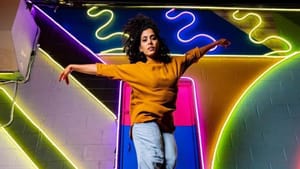 Pérez-Vega portrait shot, she’s mid-air mid-step, looking towards low-angle cam, a rainbow painted wall with neon behind her