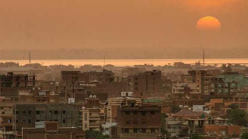 Low brown buildings crowd in front of a gleaming river, in front of a dusky orange sky with a large sinking sun at right.