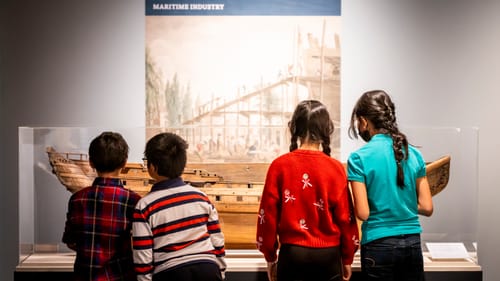 4 children, seen from behind, study an exhibit about a model ship in a museum display.