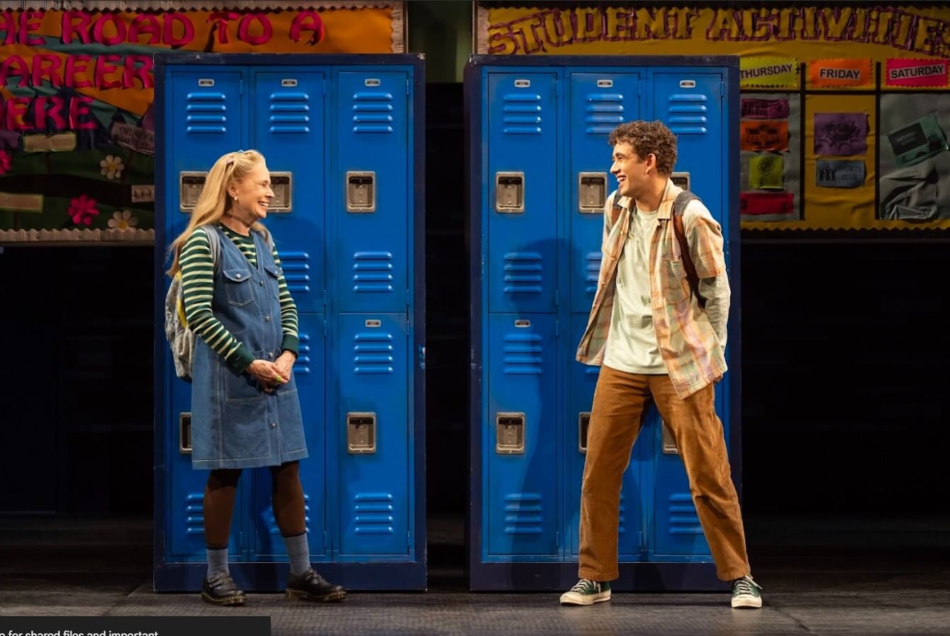 Morrison, in her late 60s but dressed as a teen, smiles shyly at Gil, a young man, in front of blue school lockers.