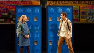 Morrison, in her late 60s but dressed as a teen, smiles shyly at Gil, a young man, in front of blue school lockers.