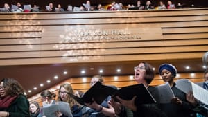 A large group singing in an indoor venue, with song books. A wall reads Verizon Hall, Home of the Philadelphia Orchestra