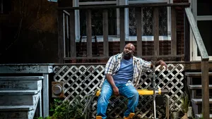 Davis, a bald, bearded Black man in an open plaid button-down, sits on a yellow work-out bench beside a decaying porch.