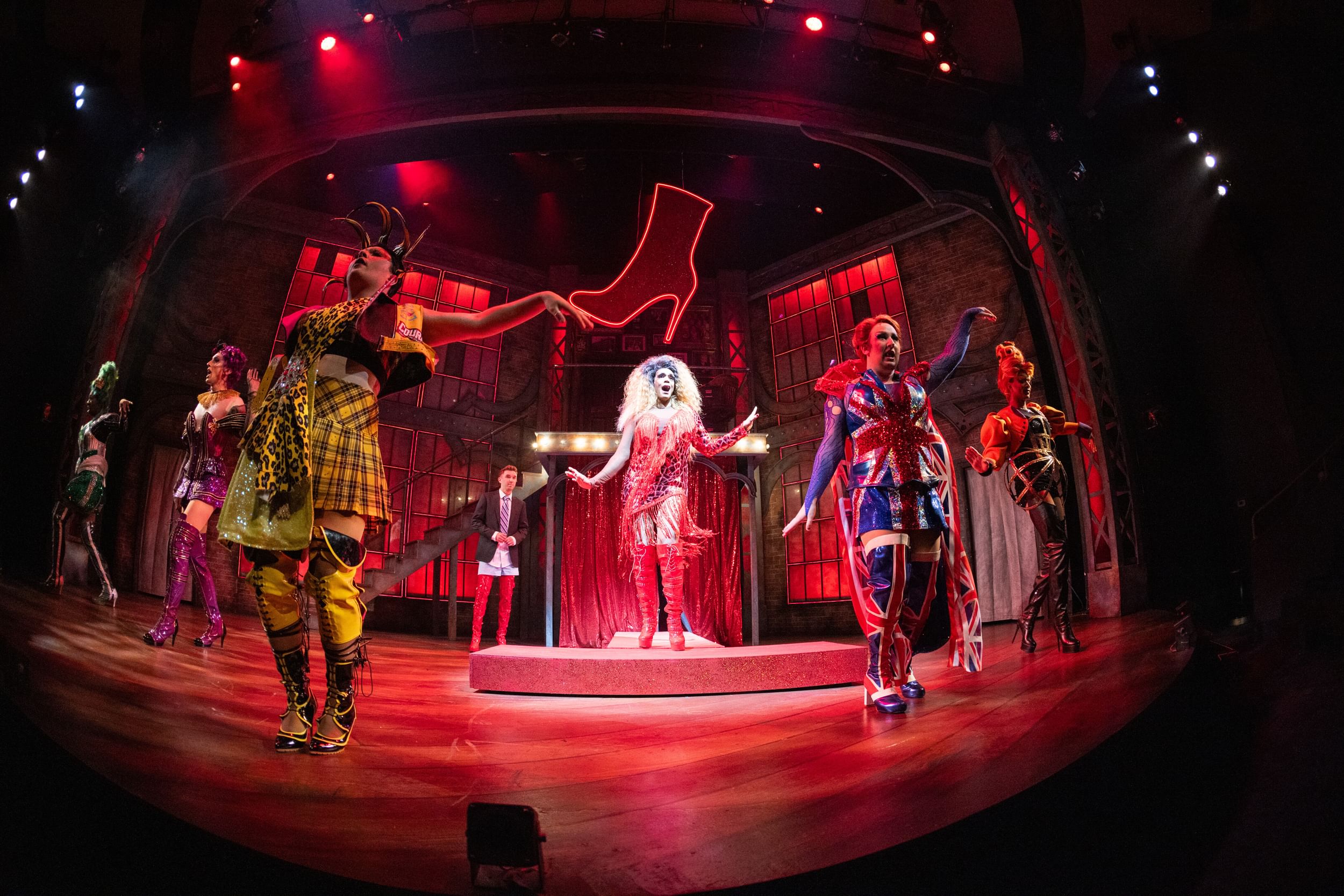 In a dramatic fish-eye lens, Diorio sings joyfully at center with 5 other actors in plaid, spangled, sparkling drag.