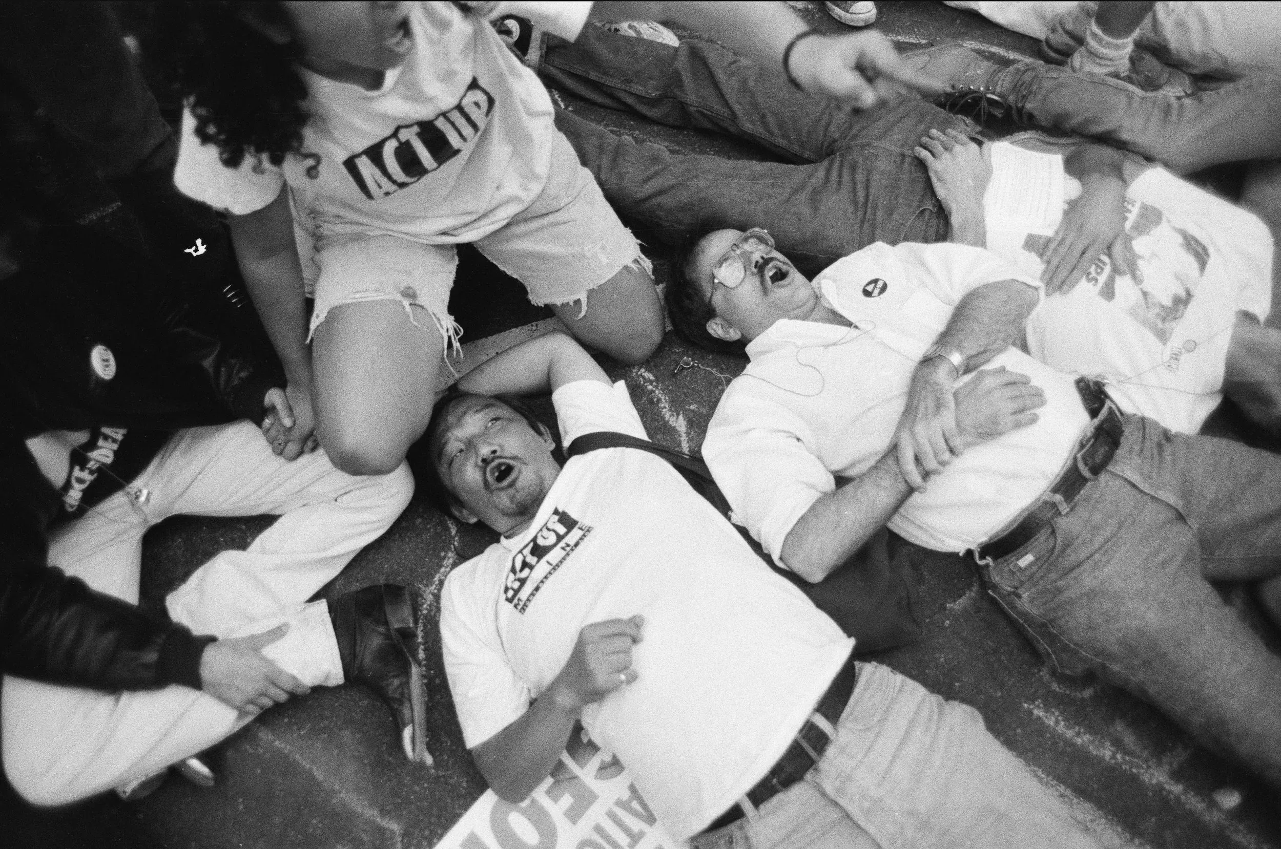 A black and white photo of people lying down, including Kiyoshi, an Asian man, wearing a white shirt with the words "Act Up."