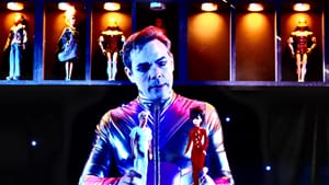 Meyer, in alluring makeup & silver bodysuit, washed in electric blue light, considers a pair of Barbie dolls he holds upright