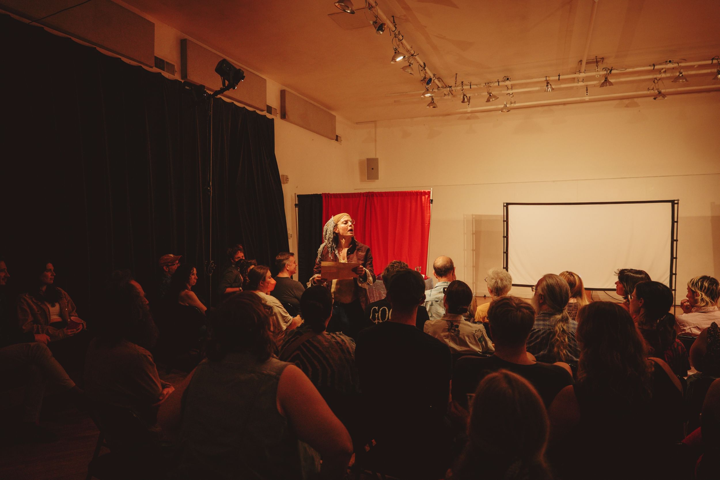 Knittel, seen over the top of a packed audience, reads from a parchment, wearing a satirical Ben Franklin costume