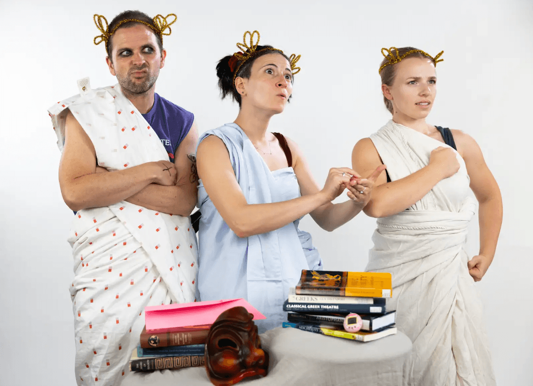 Three actors in togas made of sheets, wearing headpieces of gold pipe cleaners, strike different poses.