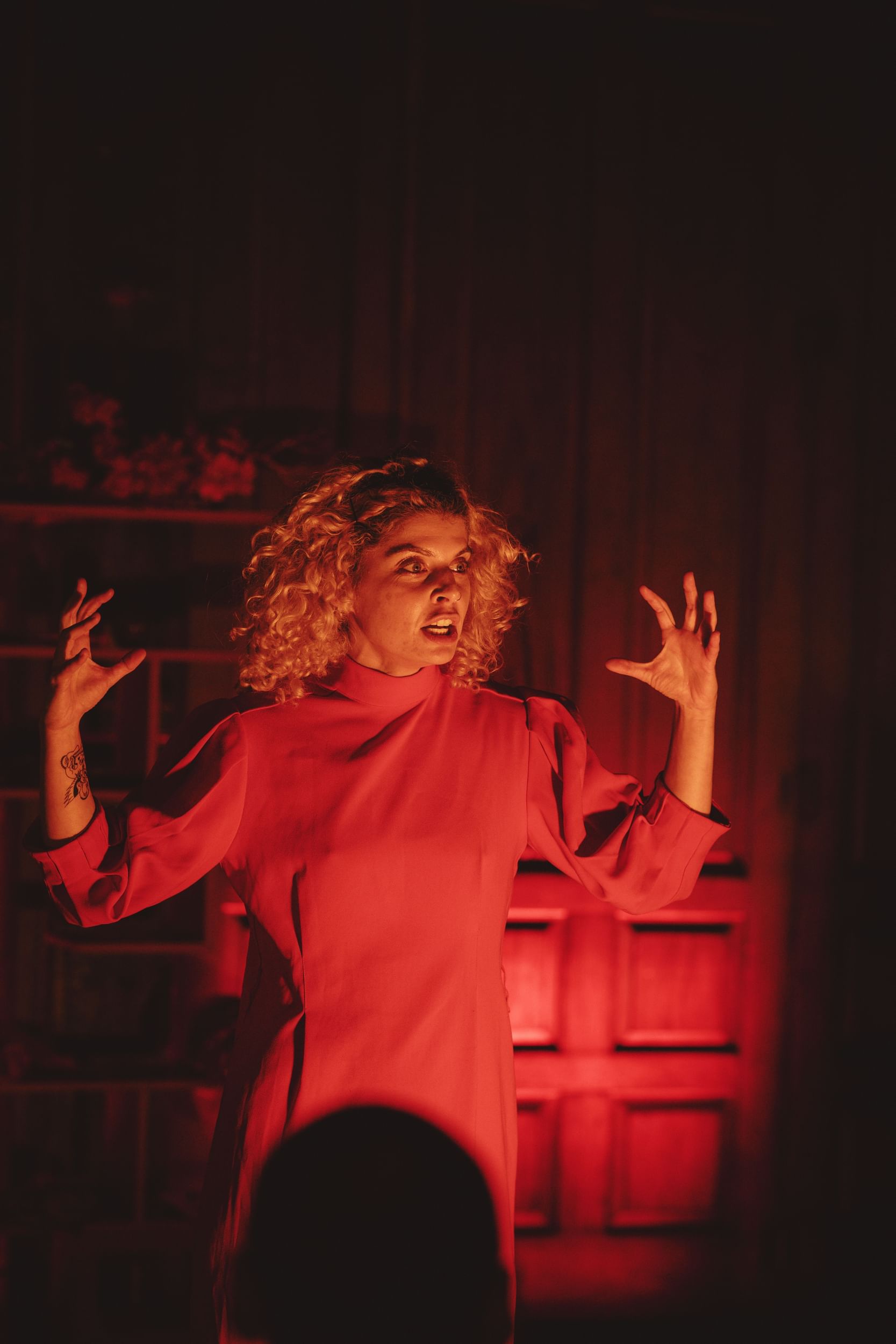 Closeup on Knittel during the show, with curly blonde hair and a baggy red dress, in a red wash of light.
