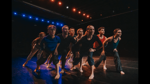 Eight dancers on stage in rehearsal, all lean forward on one leg, in mid-movement
