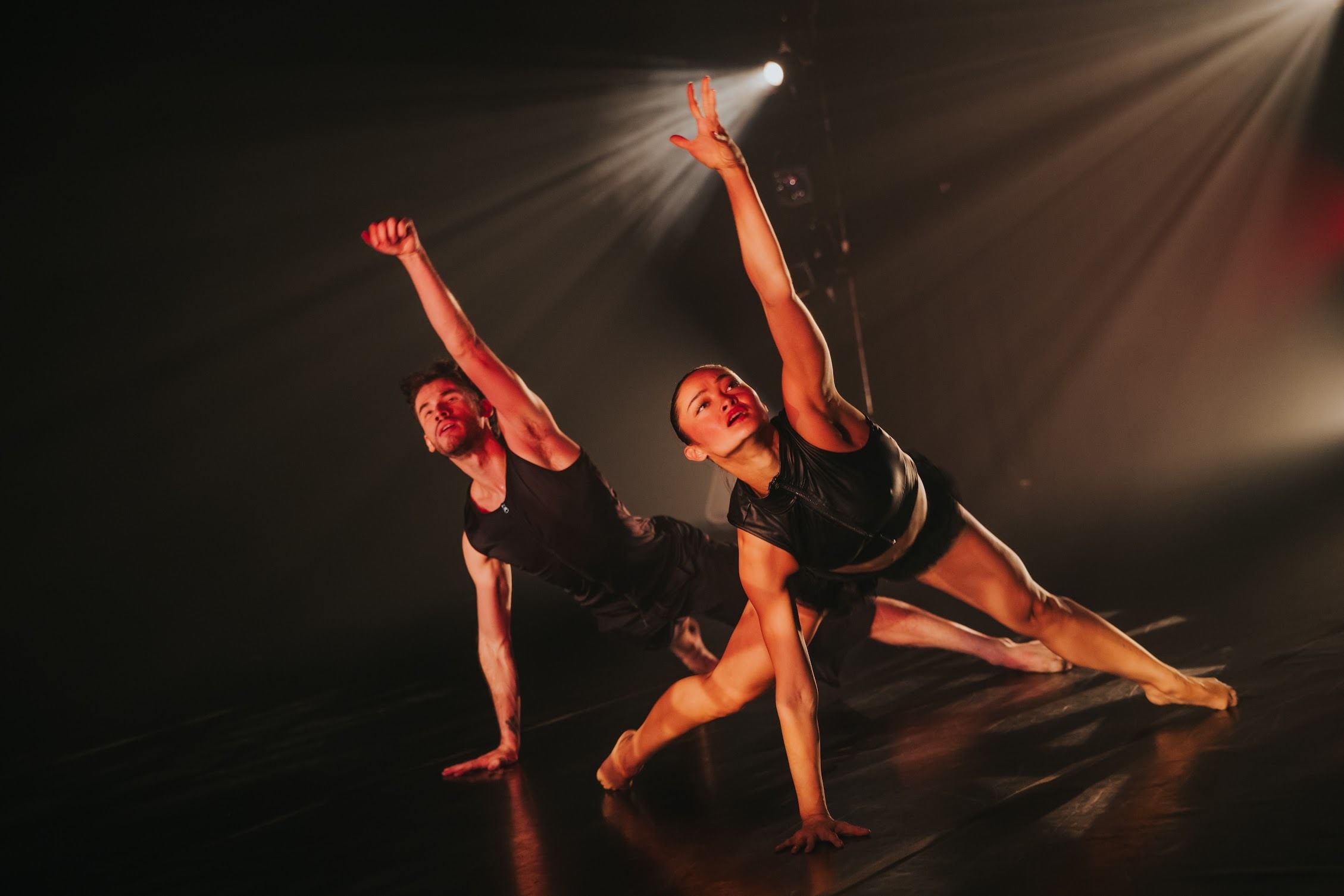 A male and a female dancer in sleeveless black costumes pose on one hand, legs split behind, one hand reaching up.