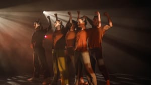 Five dancers in red shirts, loose pants stand firmly. Arms up, fists round, hard light behind them piercing dim staging