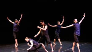 3 women pose on tiptoes with arms up; a man in black seems to control the moves of a woman dancing near the ground in front