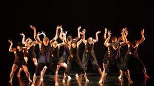 Ensemble of 10 dancers in black strike the same pose, feet set wide and arms angled inquisitively up above their heads.