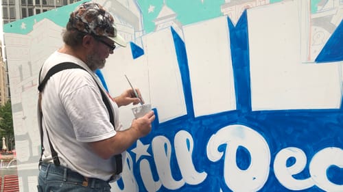Krall, in a white tee, jeans, and suspenders, dips a paintbrush into a cup while standing next to his in-progress mural.