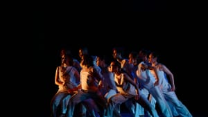 A tightly packed group of 10 dancers in white all surge to one side with a wide stance, faces turned toward an orange light.