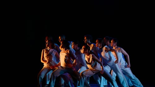 A tightly packed group of 10 dancers in white all surge to one side with a wide stance, faces turned toward an orange light.