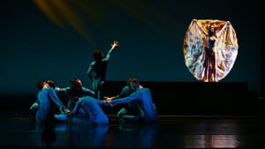 A circle of 6 dancers in blue on the floor and one standing, reaching to another whose silky orange costume falls in a circle