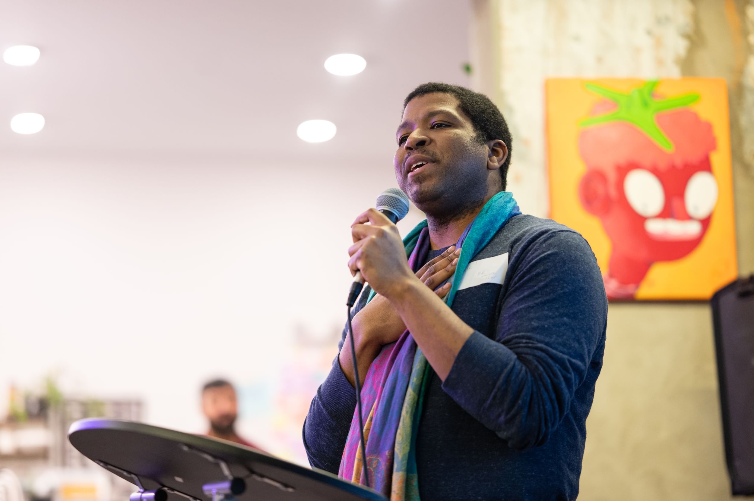 Kyle, in a blue shirt and colorful scarf, speaks into a mic with one hand on his chest.