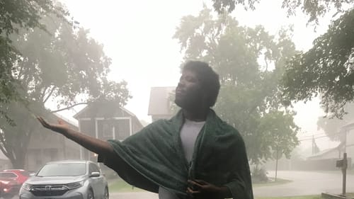 Kyle, a Black man, stands outside in the pouring rain, his arm extended, his palm open and head tilted, embracing the storm