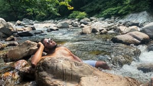 Kyle, a Black man, lies against large rocks in a river, looking up to the sunny sky while wearing sunglasses and swim trunks