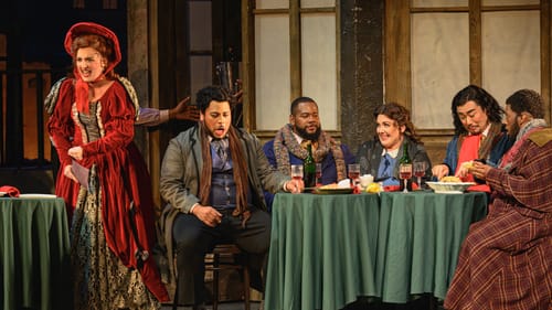 Margevich sings at left in an opulent red dress; the others sit with various expressions around green tables with wineglasses