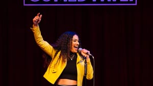 In colorful pants and a bright yellow jacket, Merci performs stand-up comedy in her role, the mic in one hand.