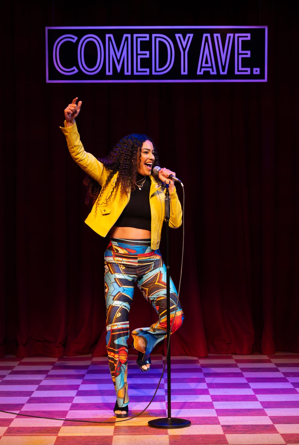 In colorful pants and a bright yellow jacket, Merci performs stand-up comedy in her role, the mic in one hand.