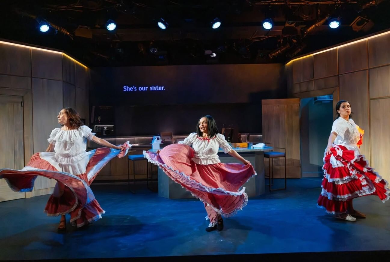 Three women, in white blouses and layered red & white ruffled skirts, twirl onstage. A caption above reads “she’s our sister”