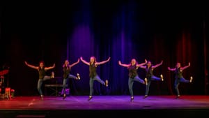 Six performers in mid-step on stage, their left legs parallel to the floor, right legs hover off the floor pointing down