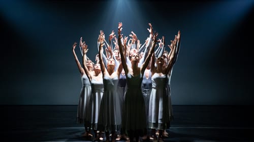 About 15 dancers in gray costumes stand together in a spotlight, arms reaching upward as if in supplication.
