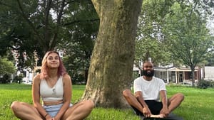 Two people sit cross-legged in the grass by a tree during the day, looking peaceful